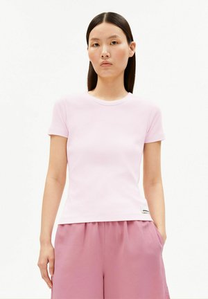 Femme aux longs cheveux noirs portant un t-shirt rose clair à manches courtes et un pantalon large rose, debout devant un fond blanc uni.
