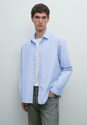 Junger Mann in einem hellblau gestreiften Hemd über einem weißen T-Shirt mit grauen Jeans, der an einer schlichten hellen Wand steht.