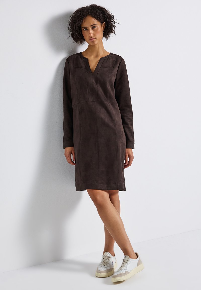 Bruine suède jurk met lange mouwen, V-hals en naaddetails, vallend boven de knie. Gepaarde met witte en beige sneakers.