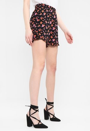 Vrouw draagt zwarte korte broek met bloemenprint en gerimpelde zoom, gecombineerd met zwarte hoge hakken met veters, staand tegen een witte achtergrond.
