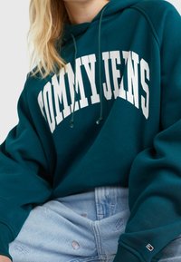 Felpa verde smeraldo con scritta bianca "TOMMY JEANS", cappuccio con lacci e polsini a coste. Abbinata a pantaloni corti in denim blu chiaro.