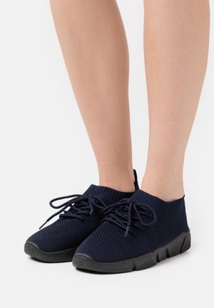 Piernas con zapatillas negras de punto con cordones y suelas gruesas de goma sobre un fondo blanco.