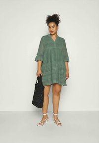 Vestido tunic verde em renda com decote em V, mangas três quartos e forma evasê. Combinado com sandálias brancas de tiras e uma mala de mão preta.