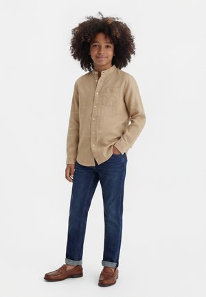 Παιδί με σγουρά μαλλιά στέκεται, φορώντας μπεζ πουκάμισο με κουμπιά, μπλε τζιν με γυριστό τελείωμα και καφέ μοκασίνια, χαμογελώντας με το ένα χέρι στην τσέπη.