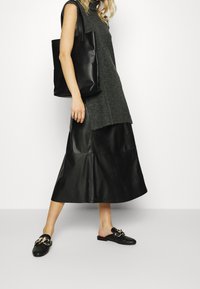 Sac fourre-tout en cuir noir, haut sans manches gris en tricot, et jupe en cuir noir avec ourlet évasé. Chaussures noires à enfiler avec des accents métalliques dorés.