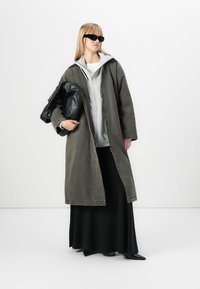 Donna con occhiali da sole, lungo cappotto grigio su una felpa grigia, gonna nera, che tiene una grande pochette di pelle nera contro uno sfondo bianco.