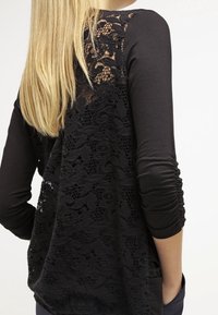 Haut noir à manches longues avec un panneau arrière en dentelle à motifs floraux. L'avant est lisse, tandis que les manches présentent des détails froncés.