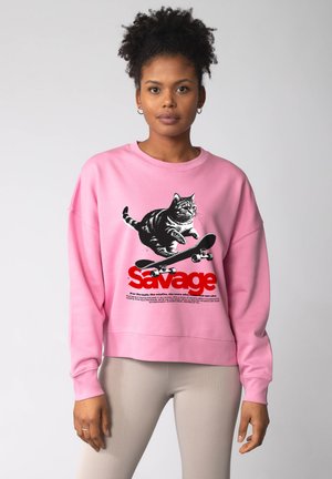 Femme portant un sweat-shirt rose avec un motif de chat noir et blanc sautant par-dessus un skateboard, accompagné du mot "Savage" en texte rouge.