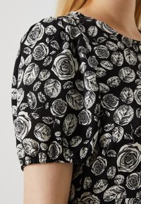 Černá látka s vzorem bílých růží a listů; má balónové krátké rukávy a kulatý výstřih; hladká textura.