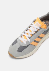 Scarpa da ginnastica in suede grigia con dettagli arancioni, lacci bianchi e tre strisce nere. Presenta una linguetta testurizzata e una suola in gomma.