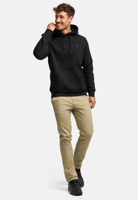 Ung mand smilende iført en sort hættetrøje, beige bukser og sorte sneakers, stående mod en lys baggrund.