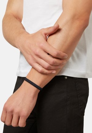 Zwarte gevlochten armband gedragen om een pols, die complementair is aan een wit T-shirt en zwarte broek. Focus op de textuur en het patroon van de armband.