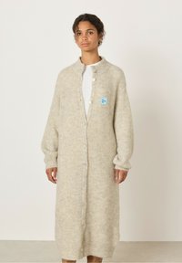 Svetlo bež pletena jakna z rebrasto teksturo, dolžina do tal, zapiranje na gumbe in majhna modra oznaka na levi strani.