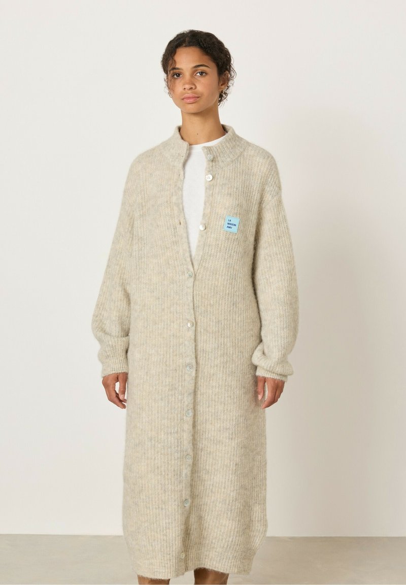 Svetlo bež pletena jakna z rebrasto teksturo, dolžina do tal, zapiranje na gumbe in majhna modra oznaka na levi strani.