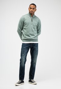 Ritssluiting, longsleeve trui in lichtgroene gebreide stof, gecombineerd met donkerblauwe jeans en zwarte sneakers. Opvallend door de kraag en het logo-detail.