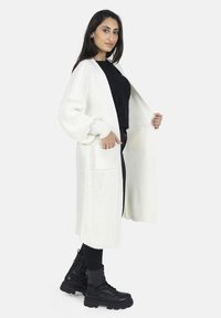 Cardigan blanc en tricot avec poches, devant ouvert, texture côtelée. Associé à un haut noir et des bottes de combat noires.