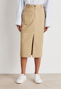 Beige Baumwollrock in knielanger Form mit einem vorderen Schlitz, Knopfverschluss und seitlichen Taschen, kombiniert mit weißen Sneakers vor einem neutralen Hintergrund.