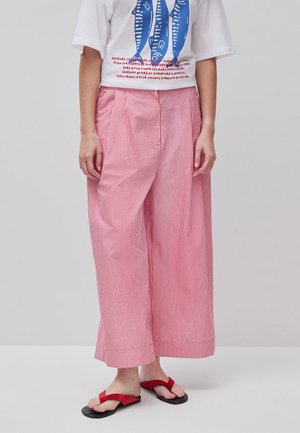 Persoon die rode en witte verticale gestreepte wijde broek draagt, wit T-shirt met een blauw vismotief en rode sandalen met bandjes.