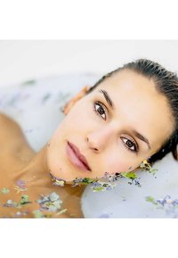 Un primo piano di un volto femminile immerso nell'acqua, circondato da piccoli petali di fiori viola e verdi, con un'illuminazione soffusa e una texture della pelle liscia.