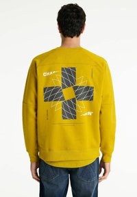 Sudadera amarilla con un patrón geométrico negro en la parte posterior. Presenta puños y dobladillo acanalados, con texto impreso sobre el diseño. Corte casual.