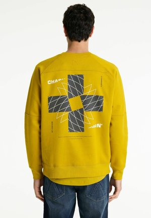 Gelbes Sweatshirt mit schwarzem geometrischem Muster auf der Rückseite. Mit gerippten Ärmelbündchen und Saum, sowie bedrucktem Text über dem Design. Lässige Passform.