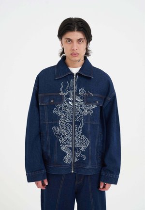 Jeansjacke in blauer Farbe mit einer weißen Drachenbestickung auf der Vorderseite und großen aufgesetzten Taschen. Mit Reißverschluss und lässigem Schnitt.