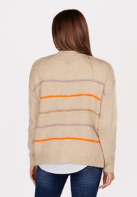 Donna con lunghi capelli castani che indossa un maglione di lana beige con righe orizzontali viola, arancioni e marroni, jeans scuri e una camicia bianca sotto.