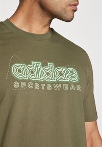 T-shirt in cotone verde oliva con logo "adidas" grande e delineato in bianco e verde, con la scritta "SPORTSWEAR" stampata sotto in bianco. Maniche corte.