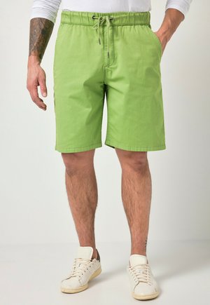 Mann trägt hellgrüne Shorts mit Kordelzug, weißes langärmeliges Hemd und weiße Sneakers, steht mit einer Hand in der Tasche.