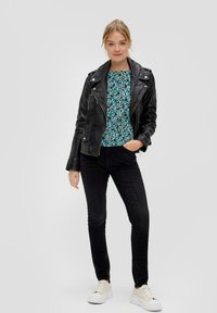 Schwarze Leder-Motorradjacke über einem bunten, floral gemusterten Oberteil, kombiniert mit schwarzen Skinny-Jeans und weißen Sneakers. Glatte Textur, taillierter Schnitt.