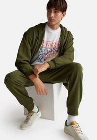 Tuta verde realizzata in materiale morbido con cappuccio zip-up e polsini elastici. La maglietta bianca presenta un testo colorato. Sneakers beige con dettagli verdi.