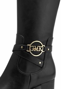 Black leather knee-high boot featuring a gold circular buckle with "GMK," a cross-strap design, and a block heel.