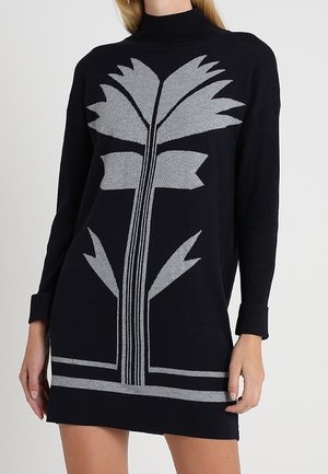 Mujer que lleva un vestido de punto negro de manga larga, con cuello alto y un gran estampado floral geométrico gris en la parte delantera.