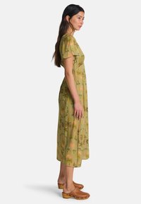 Ροή πράσινου φορέματος με λουλουδάτο σχέδιο σε πορτοκαλί και καφέ, κοντά μανίκια και άνετη εφαρμογή, συνδυασμένο με καφέ τσόκαρα.