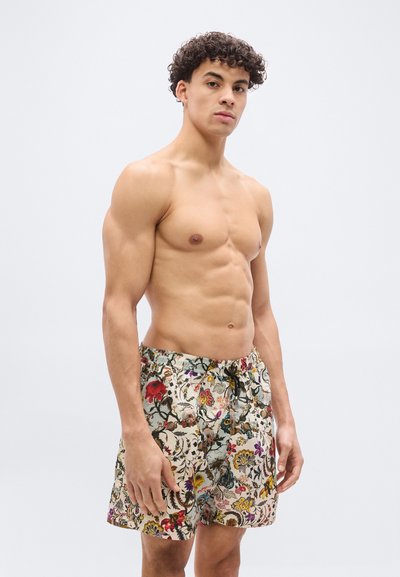 Νεαρός άνδρας με σγουρά μαλλιά που φοράει μαγιό με λουλουδάτο σχέδιο, στέκεται μπροστά σε ένα απλό ανοιχτό γκρι φόντο.