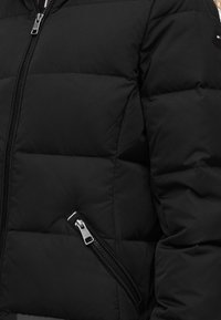 Veste matelassée noire avec fermetures éclair visibles sur les poches avant et diagonales, montrant un gros plan du tissu rembourré et des détails de couture.