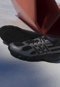 Par sorte og grå atletiksneakers med mesh og syntetiske detaljer hvilende på en betongulv nær en rød geometrisk struktur.