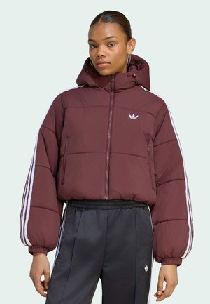 Jonge vrouw draagt een bordeauxrode gewatteerde jas met capuchon en witte strepen op de mouwen, gecombineerd met zwarte Adidas trainingsbroek met witte strepen.