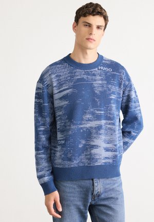 Young man standing wearing blue patterned sweater with "HUGO" logo and blue jeans against a plain white background.