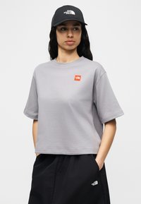 Γυναίκα που φοράει γκρι κοντομάνικο μπλουζάκι με μικρό πορτοκαλί λογότυπο, μαύρο παντελόνι με λευκό λογότυπο και μαύρο καπέλο με λευκό λογότυπο.