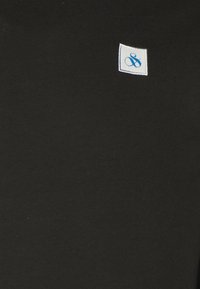 Camiseta de algodón negra con un pequeño parche cuadrado blanco que tiene texto en azul. La tela tiene una textura suave y ligeramente elástica.