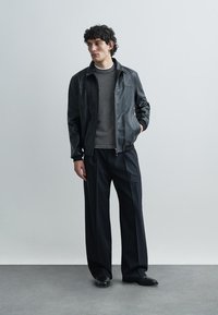 Černá kožená bunda, šedý svetr s pleteným vzorem a široké černé kalhoty. Model stojí před světle šedým pozadím.