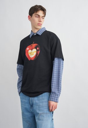 Joven con camisa de cuadros azules superpuesta y camiseta negra con gráfico de manzana roja, y jeans desgastados, mirando hacia su izquierda.