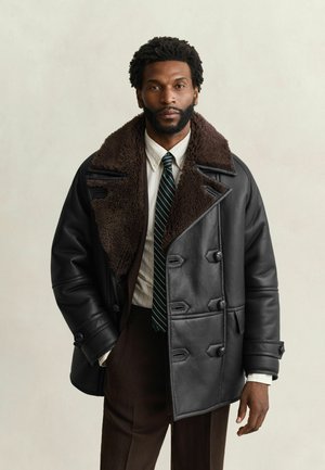 Man draagt een zwarte leren shearling jas, wit overhemd, groene gestreepte stropdas en donkerbruine broek tegen een neutrale achtergrond.