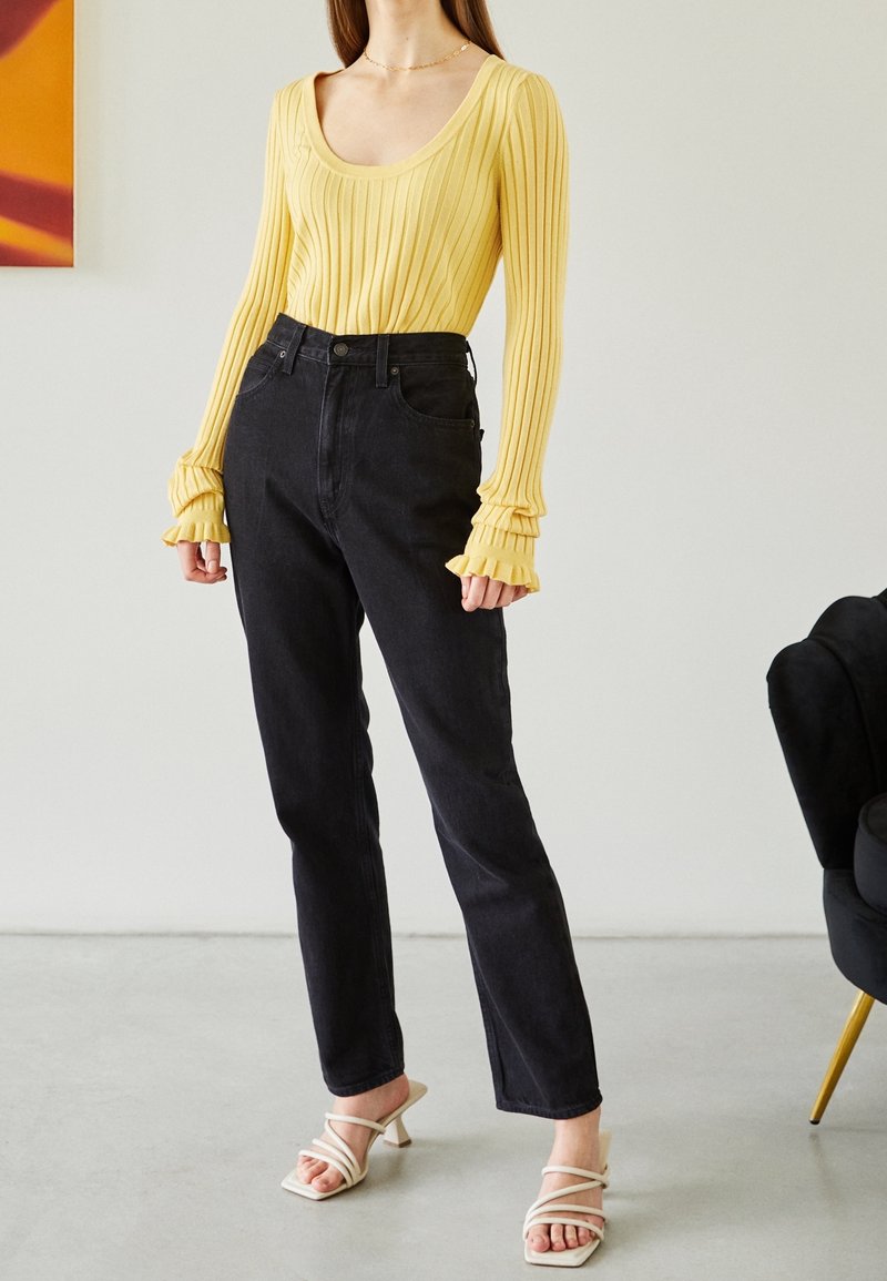 Femme portant un haut jaune côtelé à manches longues avec des poignets volantés, un jean noir taille haute et des sandales blanches à talons avec des brides.