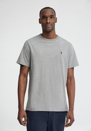 Mann trägt ein schlichtes hellgraues T-Shirt mit kurzen Ärmeln und kleinem dunklem Logo auf der linken Brust, kombiniert mit dunkler Hose, vor einfarbigem Hintergrund stehend.