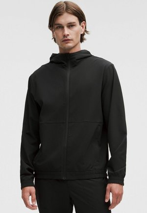 Črna jopa s kapuco z teksturirano površino, z zadrgo spredaj in elastičnimi manšetami. Vsebuje dva stranska žepa in je lahka, zračna zasnova.