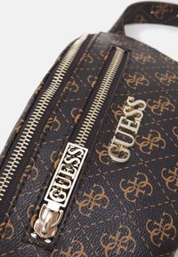 Brązowa syntetyczna torba z tłoczonym wzorem logo, złotymi okuciami i zamkiem błyskawicznym. Zawiera wyraźny detal ozdobny z napisem "GUESS".
