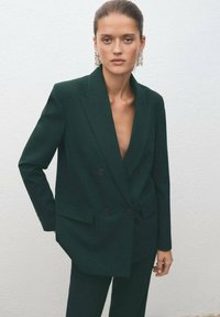 Zielona, dwurzędowa marynarka z głębokim dekoltem w kszta�łcie litery V, o strukturalnym kroju, z fakturowanej tkaniny, z czarnymi guzikami. Noszona na dopasowanych spodniach.