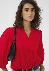 Blouse rouge légère avec un profond décolleté en V et un détail froncé. Elle possède des manches longues et est portée avec un sac bandoulière noir et un jean en denim.